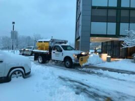 Commercial snow removal truck clearing snow from a parking lot outside a modern building, showcasing McLeod Landscaping's snow management services.