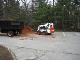 Bobcat loader unloading mulch from a truck in a residential landscaping project by McLeod Landscaping, surrounded by trees.