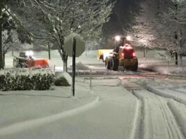 Commercial snow removal in progress at night, featuring snowplows clearing a snow-covered parking lot surrounded by trees.