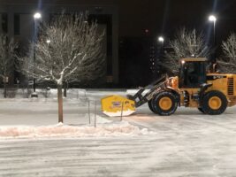 Commercial snow removal equipment clearing snow in a parking lot at night, featuring a loader with a snow plow and surrounding trees.