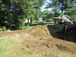 Residential landscaping project in progress, featuring a worker next to a dirt mound, with a Bobcat machine nearby, surrounded by trees and a green lawn.