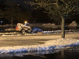 Commercial snow removal in action, featuring a snow plow clearing snow from a parking lot at night, showcasing McLeod Landscaping's snow management services.