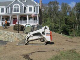 Residential landscaping project featuring a skid steer loader working on a yard in front of a large house, with landscaping materials visible in the background.