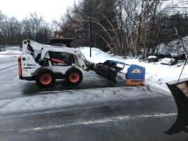 Commercial snow removal equipment in action, featuring a skid steer loader with a snow plow attachment clearing a snowy parking lot, showcasing McLeod Landscaping's snow management services.
