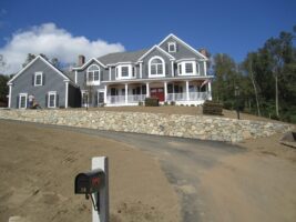 Residential landscaping project showcasing a large gray house with a stone retaining wall and a driveway, emphasizing McLeod Landscaping's design and construction work.