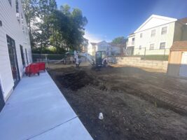 Commercial landscaping project in progress, featuring excavation equipment and workers at a site with freshly turned soil, adjacent to a newly paved walkway.