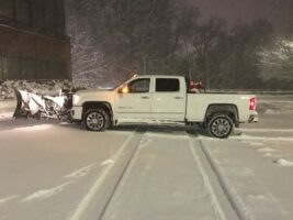 White pickup truck with snow plow clearing snow in a commercial property during a snowstorm, showcasing McLeod Landscaping's snow removal services.