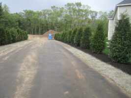 Residential landscaping project featuring a paved driveway lined with evergreen trees, mulch beds, and a construction site in the background, showcasing McLeod Landscaping's design services.