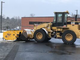 Commercial snow removal equipment, featuring a yellow snow plow attached to a CAT loader, clearing snow from a parking lot.
