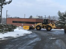 Commercial snow removal in action, showcasing a yellow snow plow clearing snow from a parking lot near a commercial building, emphasizing McLeod Landscaping's snow management services.