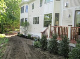 Residential landscaping project by McLeod Landscaping featuring newly planted shrubs along a dirt pathway beside a house, showcasing professional landscaping design and construction.