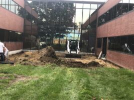 Commercial landscaping project featuring excavation work with a skid steer loader, surrounded by a grassy area and modern brick buildings.