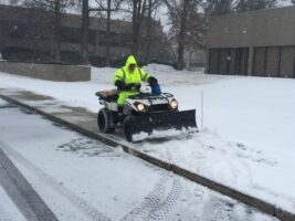 Commercial snow removal in action, featuring a worker in a bright yellow jacket operating an ATV with a snow plow, clearing snow from a sidewalk in a snowy environment.