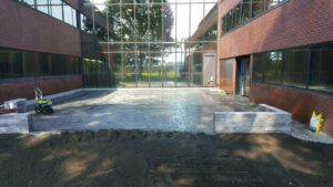 Commercial landscaping project showcasing newly installed hardscaping features, including a stone patio and retaining walls, with construction equipment visible in front of a modern commercial building.
