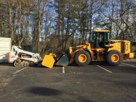 Heavy machinery for commercial snow removal, including a yellow loader and a smaller skid steer, parked in a lot surrounded by trees.