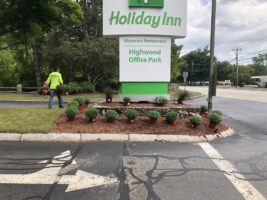 Commercial landscaping project at Holiday Inn, featuring neatly arranged shrubs and mulch around the hotel sign, with a worker in a green shirt maintaining the area.