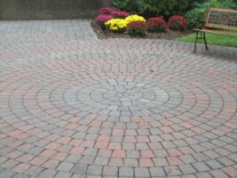 Paver stone patio installation featuring a circular pattern, surrounded by colorful flower beds, showcasing McLeod Landscaping's hardscaping services.