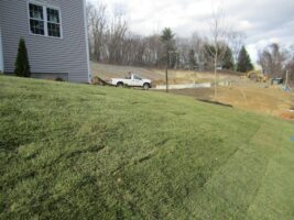 Residential landscaping project featuring freshly laid sod on a sloped lawn beside a house, with construction equipment visible in the background, showcasing McLeod Landscaping's work.