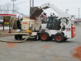 Skid steer loader operating a commercial landscaping material spreader, showcasing McLeod Landscaping's equipment for landscape installations and maintenance.