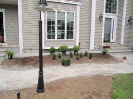 Residential landscaping project featuring newly planted shrubs and plants in front of a house, with a decorative lamp post and freshly laid stone pathway.