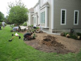 Residential landscaping project with workers planting shrubs and preparing the garden area beside a house, showcasing McLeod Landscaping's design and construction services.