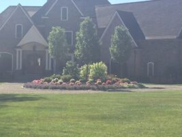 Residential landscaping project featuring a vibrant flower bed with colorful blooms, surrounded by lush greenery and a well-maintained lawn in front of a large brick home.