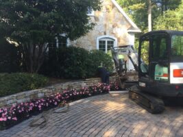 Landscaping project featuring a worker using a mini-excavator near a stone wall, surrounded by colorful flower beds and a residential home, showcasing McLeod Landscaping's design and construction services.