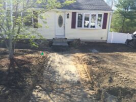 Residential landscaping project featuring a newly graded pathway leading to a house with yellow siding and maroon shutters, surrounded by fresh soil and a tree.