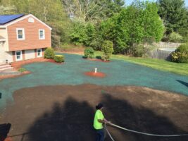 Residential landscaping project by McLeod Landscaping featuring a worker preparing a yard with green mulch and flower beds near a home.