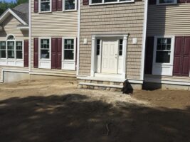 Residential landscaping project featuring a newly constructed home facade with beige siding, maroon shutters, and a front entrance, showcasing prepared ground for landscaping by McLeod Landscaping.