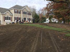 Residential landscaping project by McLeod Landscaping featuring a newly prepared lawn area in front of a house, with visible tire tracks and workers present, showcasing landscaping services.