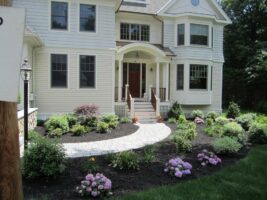 Residential landscaping design featuring a well-maintained flower bed with colorful blooms, a curved stone pathway leading to a house entrance, and a modern home with large windows and a welcoming porch.