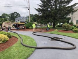 Landscaping professionals installing irrigation systems in a residential yard, featuring green grass, shrubs, and a paved driveway.