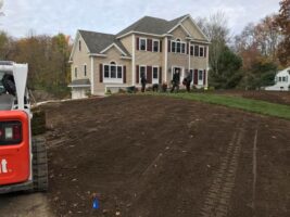 Residential landscaping project featuring a newly graded yard in front of a modern home, with landscaping equipment visible and workers preparing the area for installation.