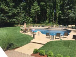 Residential landscaping project featuring a well-maintained pool area, surrounded by lush green grass, patio seating, and a serene wooded backdrop, showcasing McLeod Landscaping's expertise in outdoor design and installation.